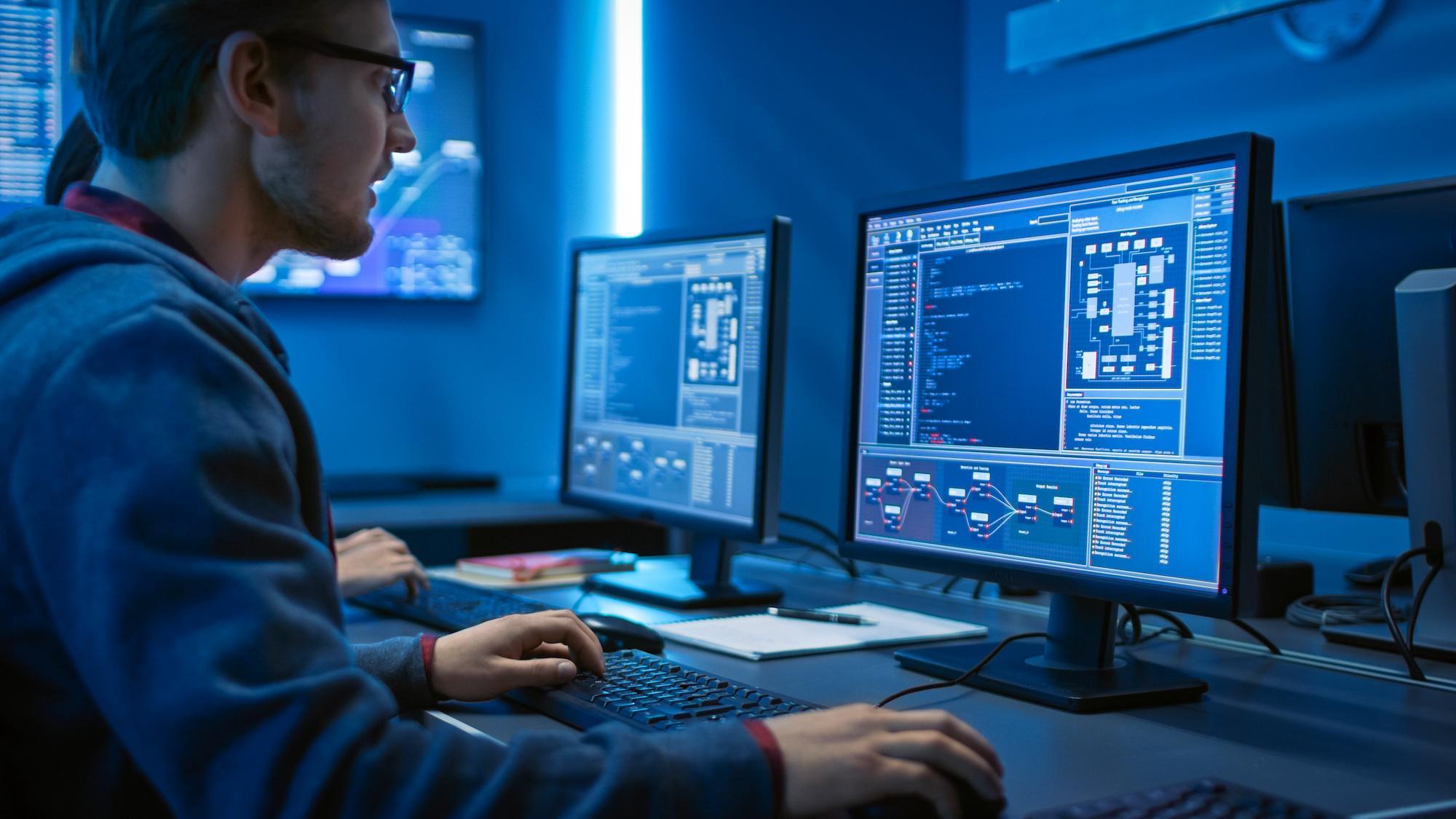 Advanced Cybersecurity MSc student working in a specialist computing lab, analysing software systems and writing code on dual monitors displaying network security tools and threat‑detection interfaces.