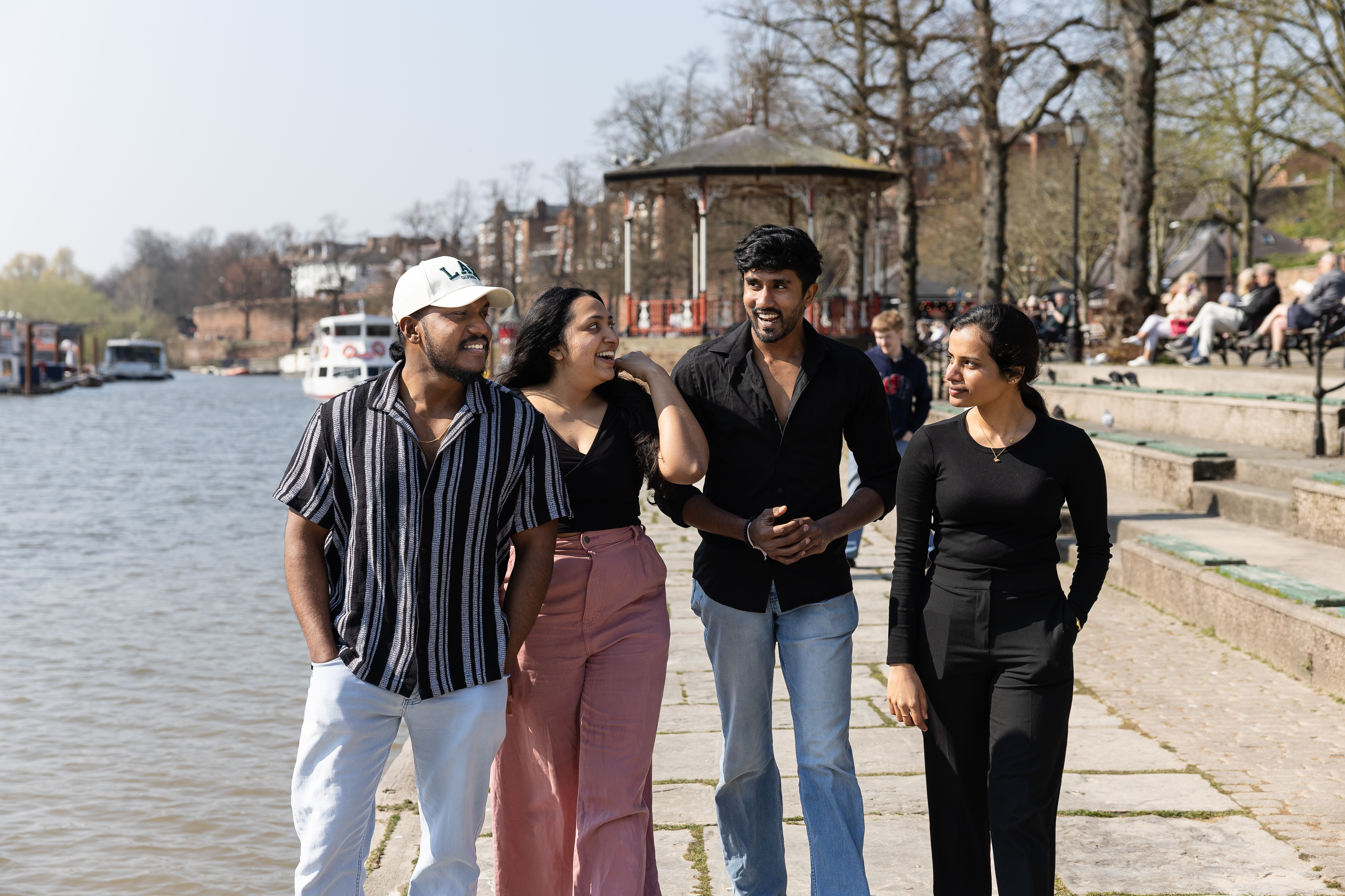 Four students walking together along a riverside promenade on a sunny day, smiling and chatting, with boats on the water and a bandstand in the background.