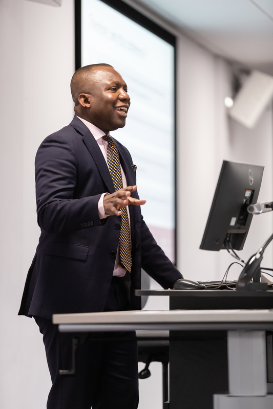 A tutor standing in front of a computer, desk and wall screen, giving a lecture in classroom.