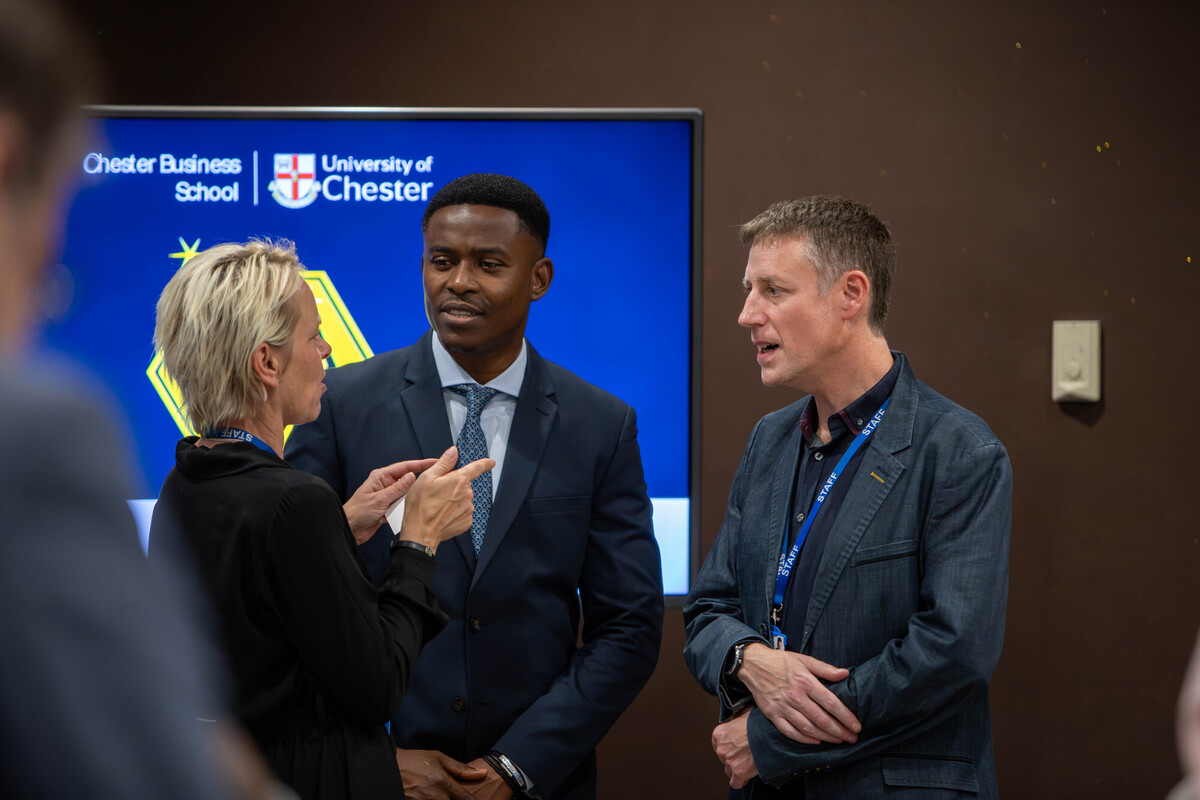 Two men and a woman speaking together at MBA award ceremony, open computer screen in the background with text for Chester Business School.