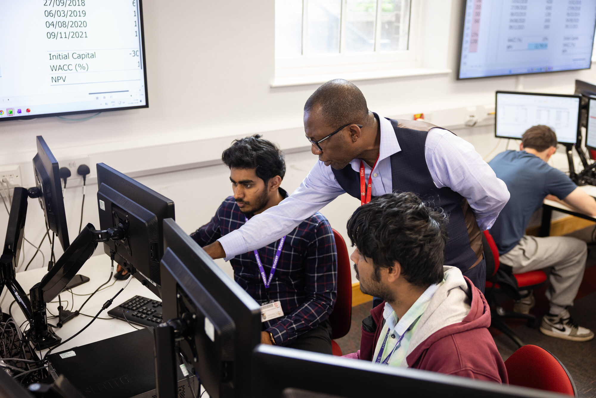 Two students sitting at computer screens, and Course Leader standing next to them, in the Fintech Suite