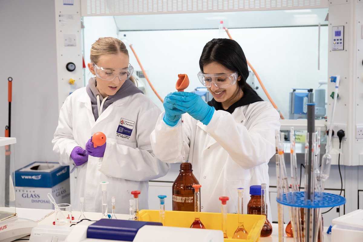 Two students working together in a laboratory wearing protective white coats and glasses.