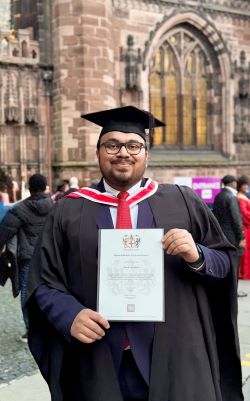Piyush standing outside Chester Cathedral in his cap and gown holding his graduation certificate