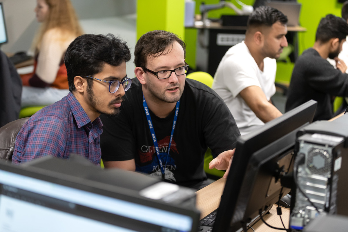 Two students sitting at desk looking at laptop while other students are studying.