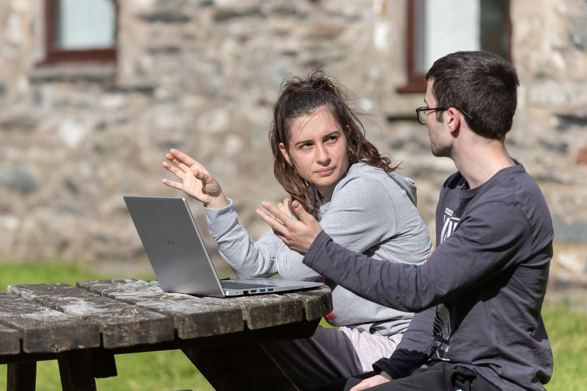 Two people sitting at a table in front of an open laptop screen in background the room has brick effect decoration.