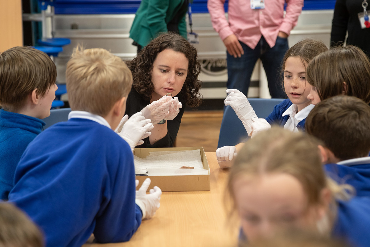 A woman wearing a blue shirt and white gloves speaks to a group of children in a classroom setting for a PGCE course.
