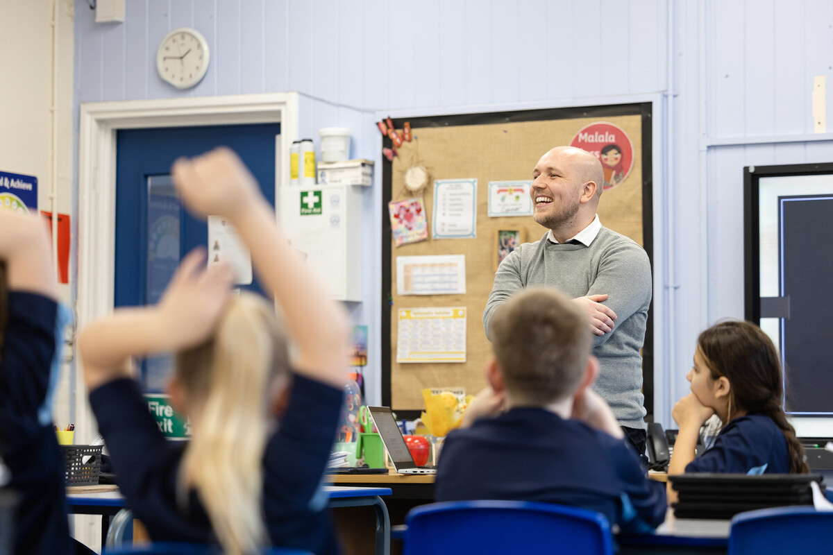 A classroom scene where an educator stands at the front near a desk, addressing a group of learners who are seated and raising their hands.