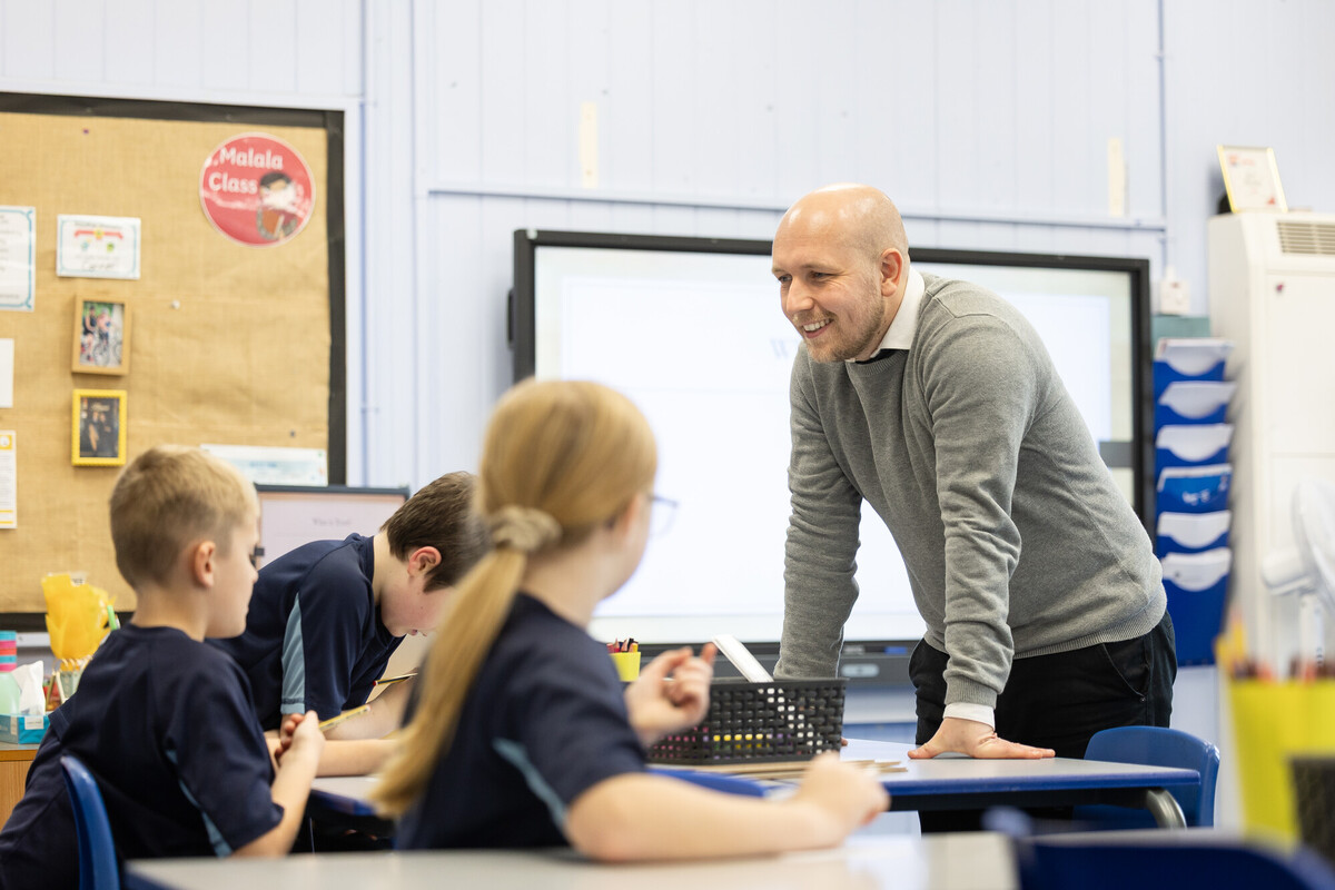 A classroom scene where an adult is leaning over a table, engaging with a small group of learners seated at desks.