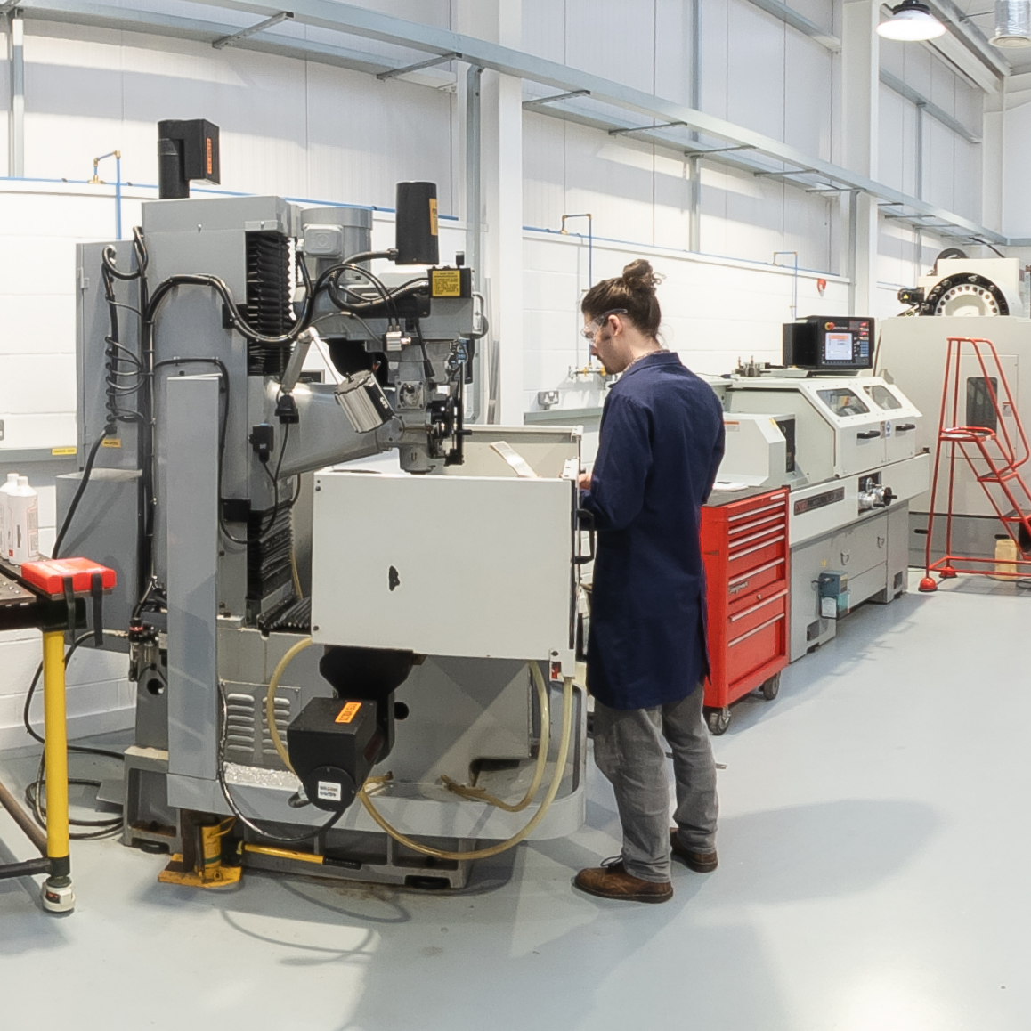 A person operates a large CNC machine in a clean, modern engineering workshop, with tools, control panels, and equipment visible around the workspace.
