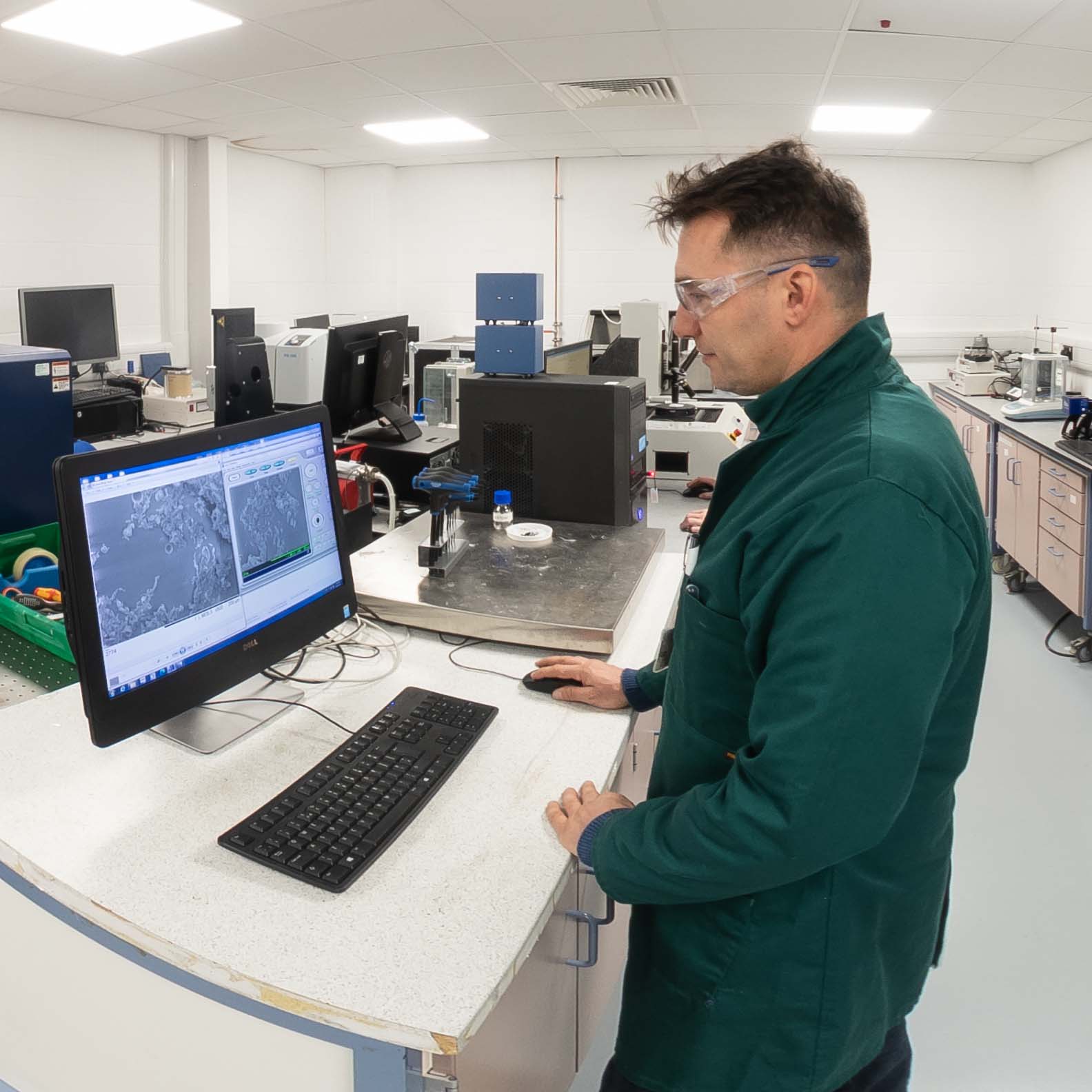 A person wearing safety goggles and a lab coat operates a computer displaying microscope images in a well-equipped laboratory filled with scientific instruments.