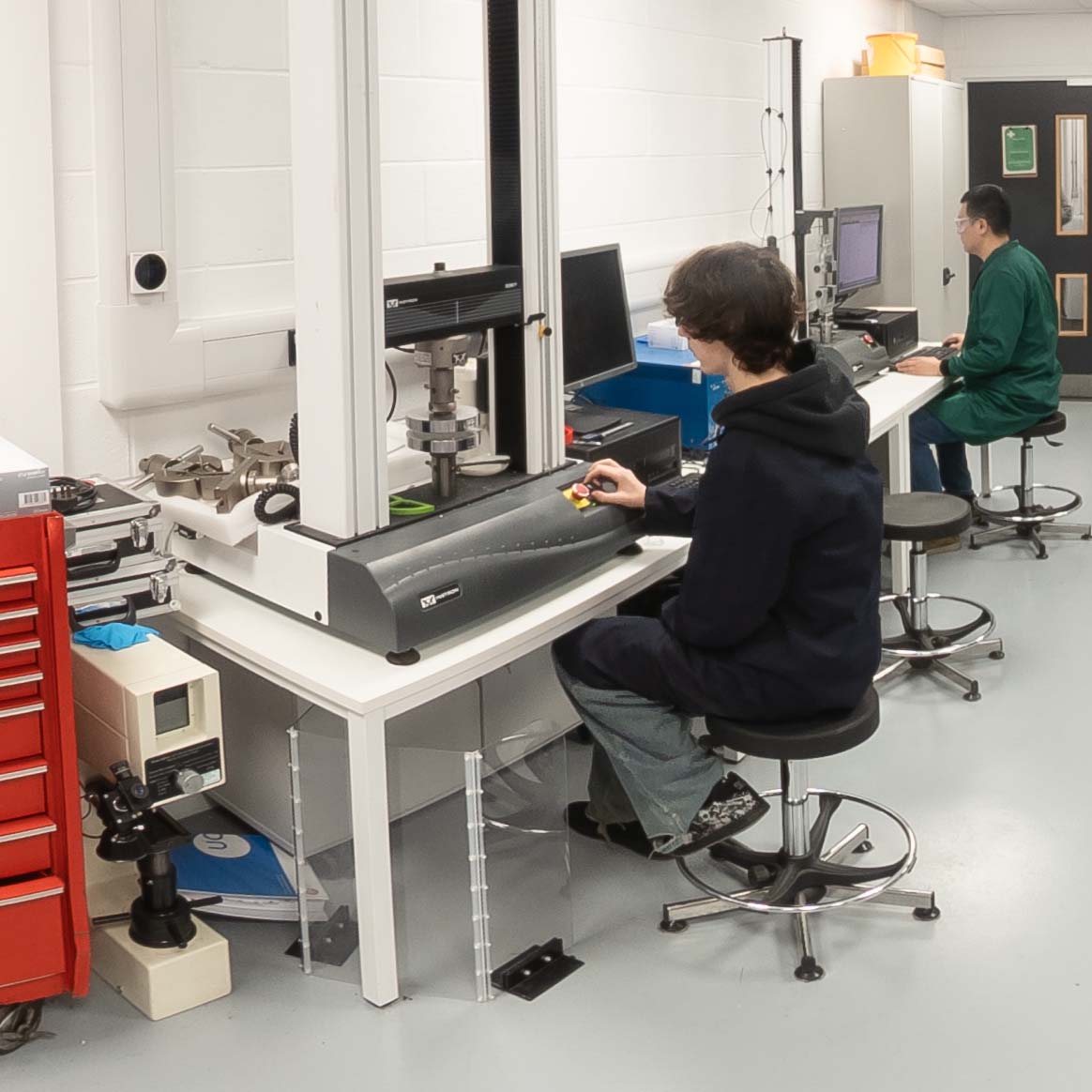 A laboratory workspace where one person operates a materials testing machine while another works at a computer station in the background.