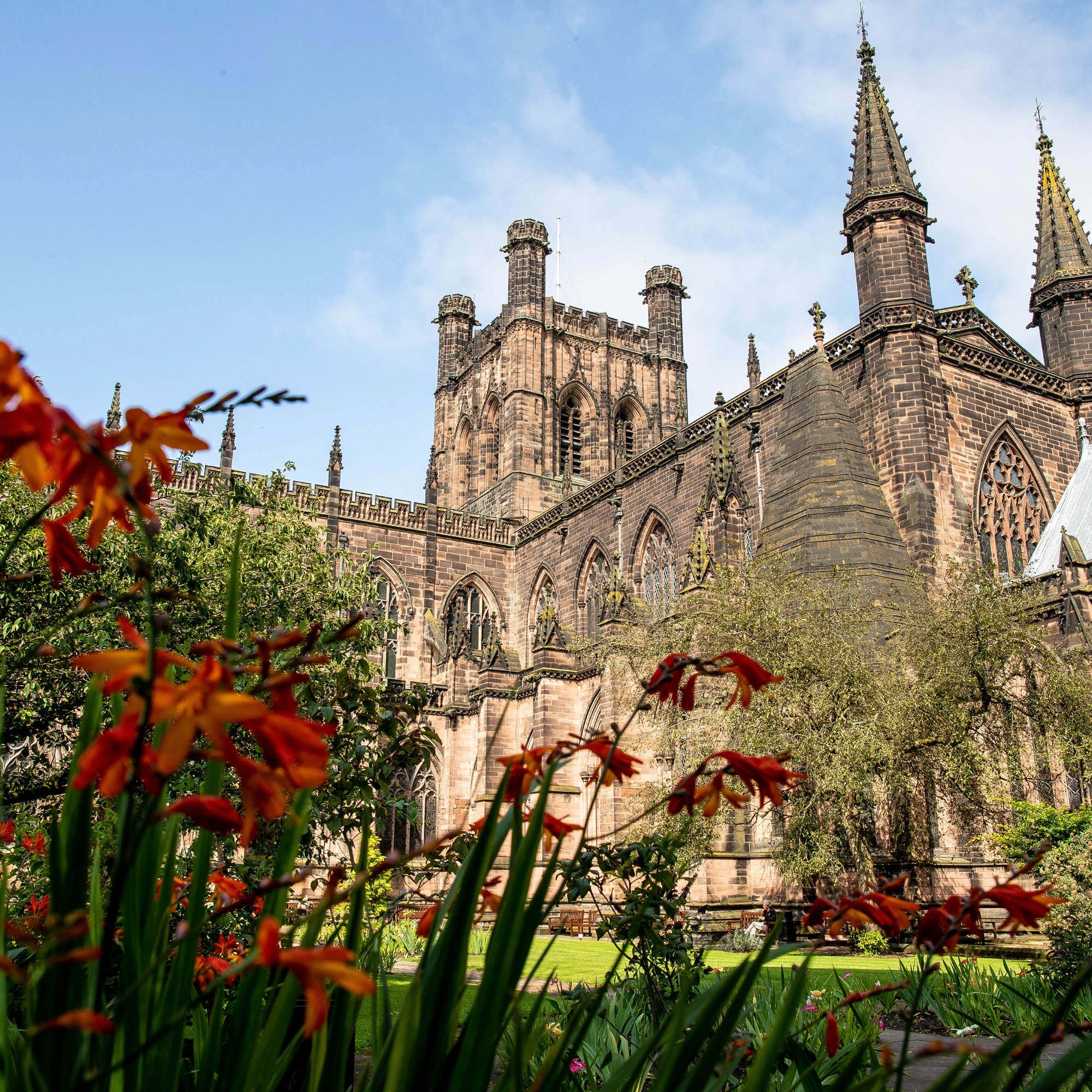 Chester Cathedral viewed from the gardens, with orange flowers in the foreground and the cathedral’s stone towers rising against a blue sky.