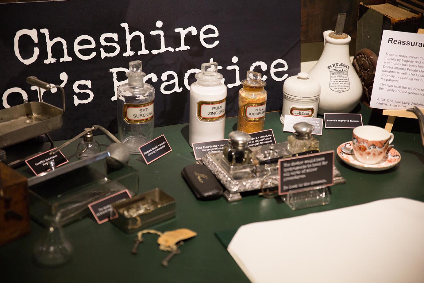 A museum-style display of Victorian-era medical and dental equipment laid out on a green tabletop, including glass bottles, jars, metal instruments, apothecary containers, and a porcelain cup and saucer.