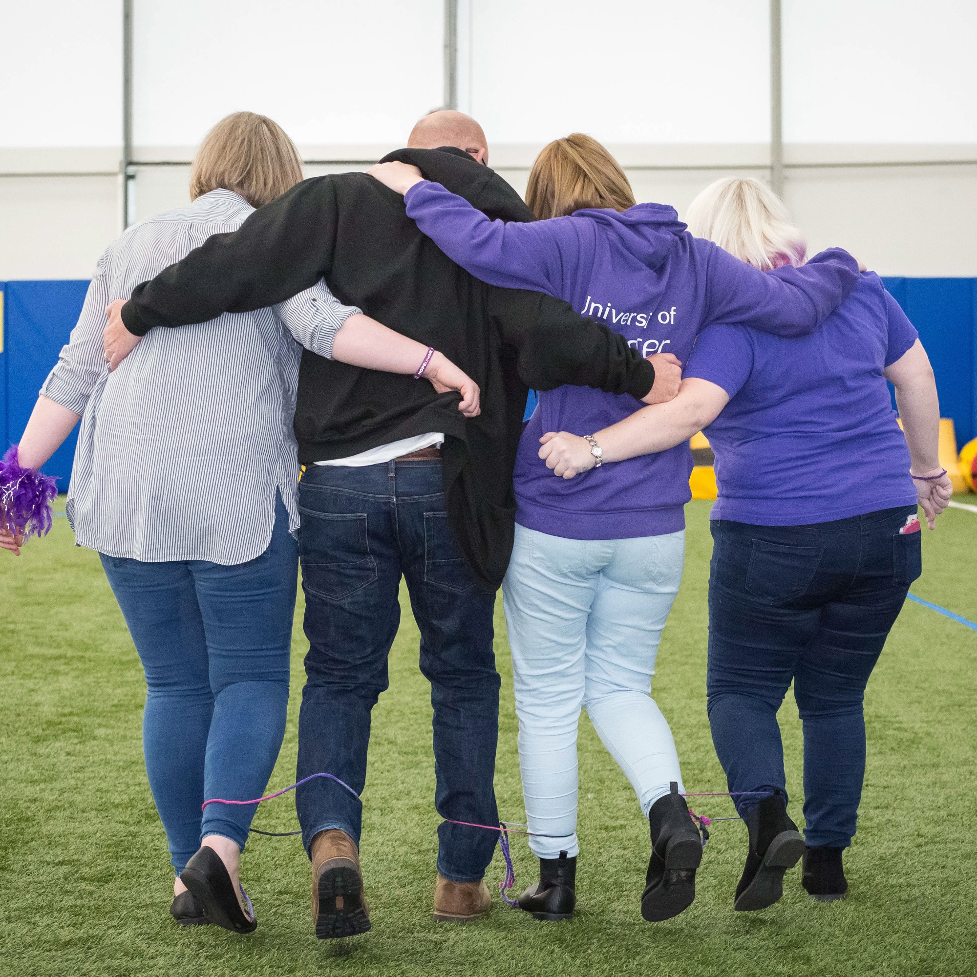 Group of participants walking arm in arm during a team-building activity on an indoor sports pitch, with their legs loosely tied together for a coordination challenge.