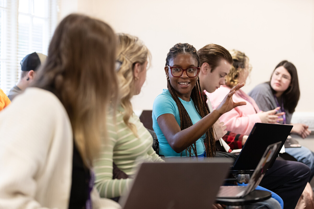 A group of diverse students sit in a classroom setting, engaged in conversation.