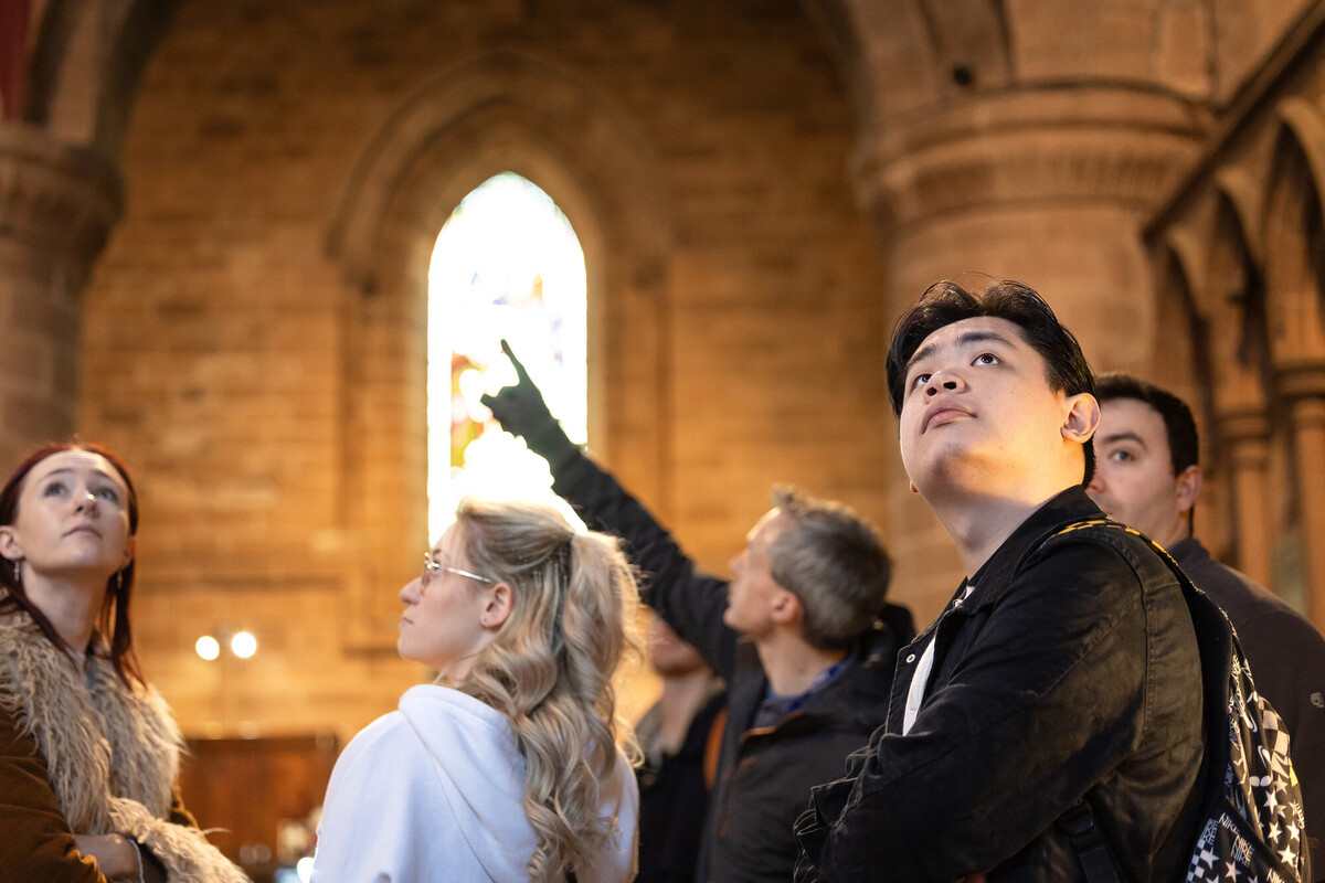 A group of students stand inside a historic stone building, looking toward a stained-glass window as someone points upward.