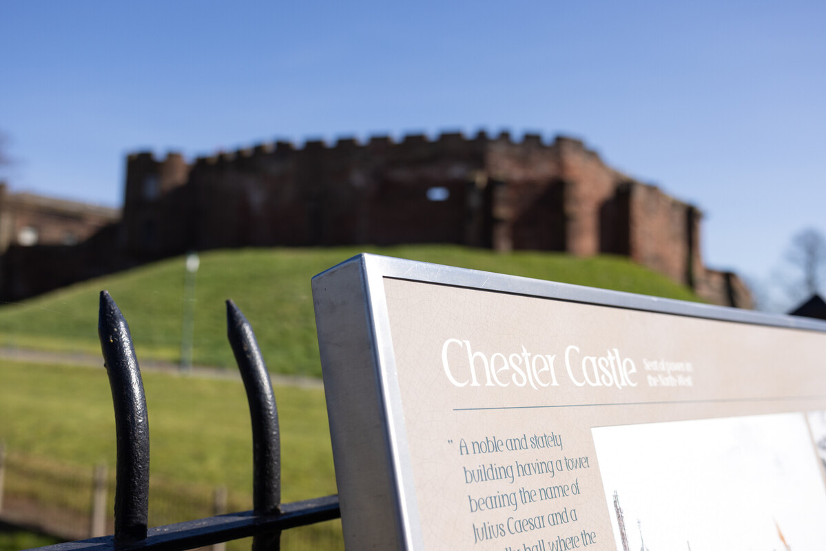 Chester Castle in Cheshire viewed from ground level, with historic stone fortress walls rising behind an informational sign, illustrating a real-world study site for History BA students exploring medieval English heritage and archaeological landscapes.