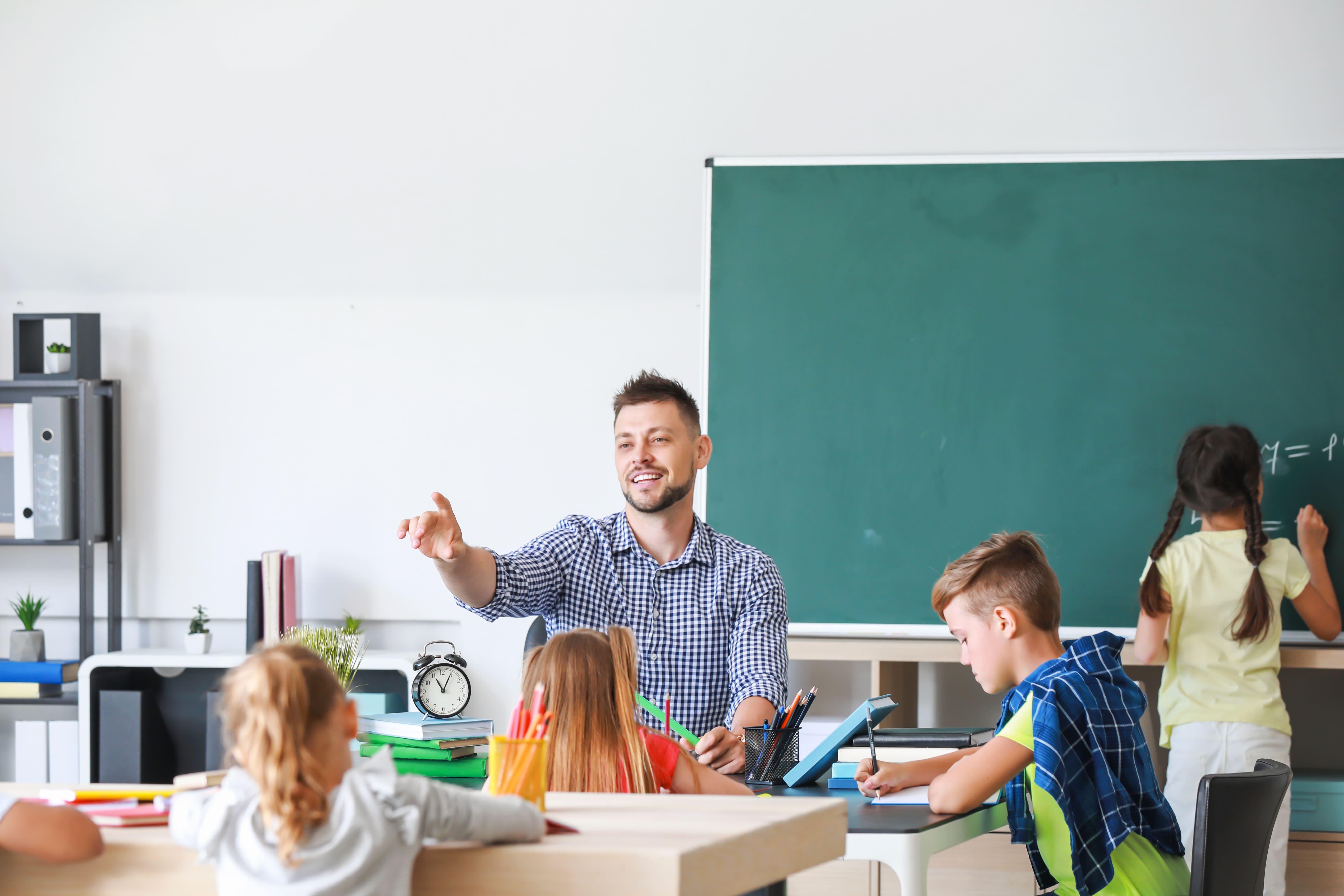 a male teacher in a classroom with childeren sitting at group tables