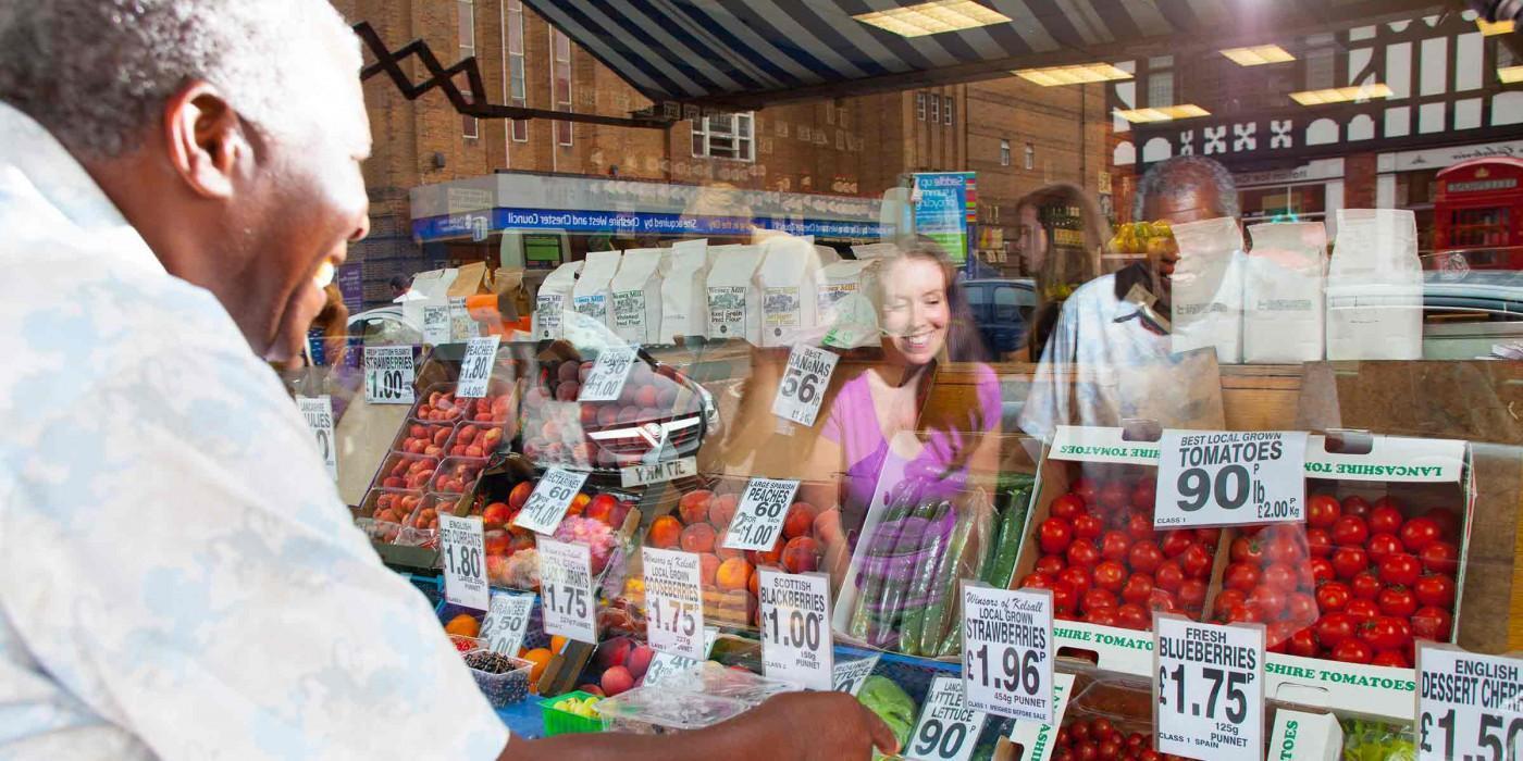 People shopping at a fresh food market with fruit and vegetables on display, illustrating community health, nutrition, and public health initiatives. Image for the Master of Public Health (MPH) degree at the University of Chester, focusing on population health, health promotion, and disease prevention.