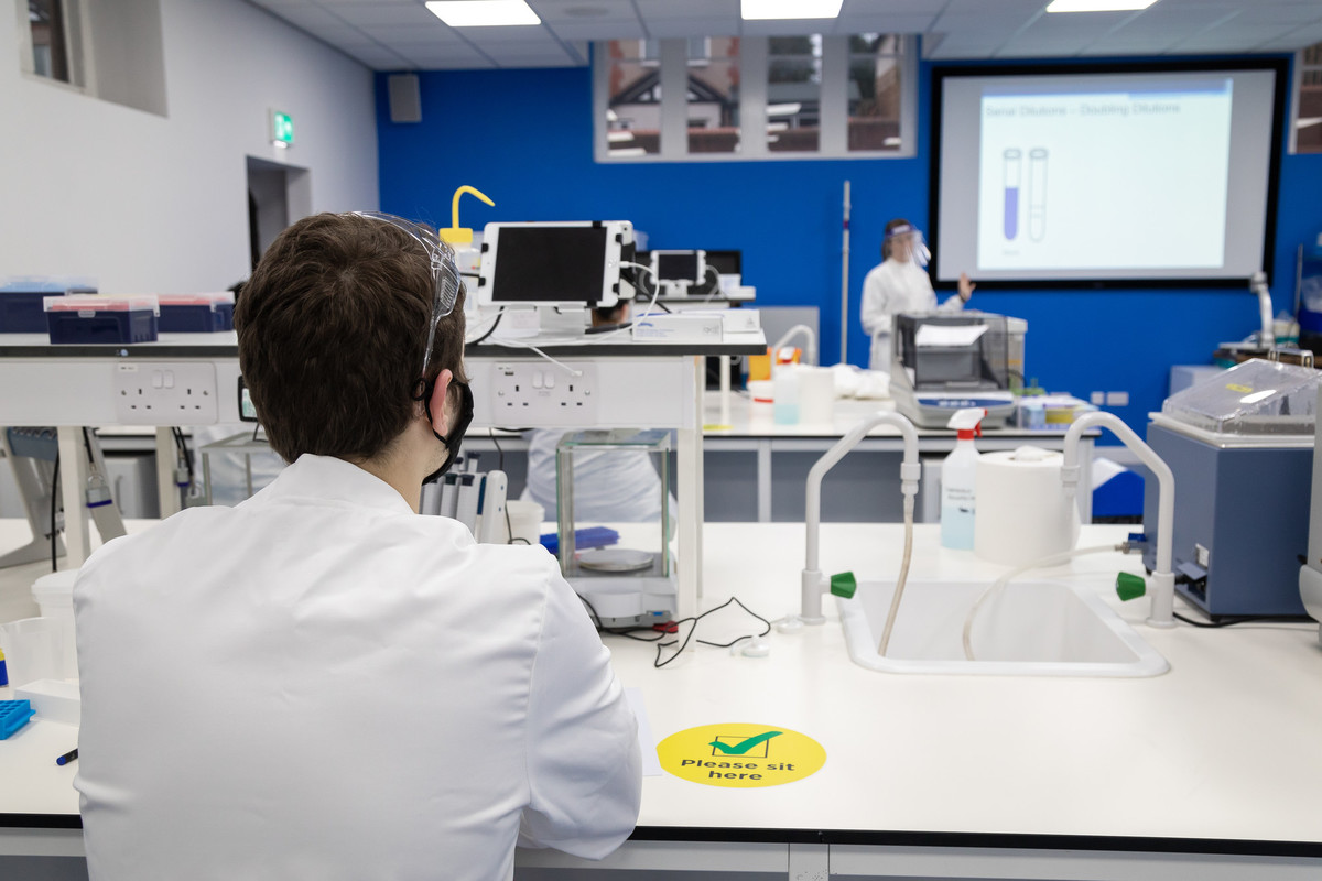 A male student working at Laboratory desk with testing equipment. A tutor is standing next to wall screen.