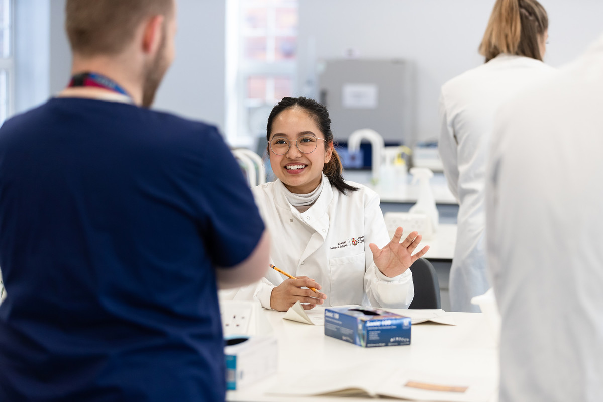 A female medical student wearing white lab coat is taking notes, and talking with male student standing in front of work desk.