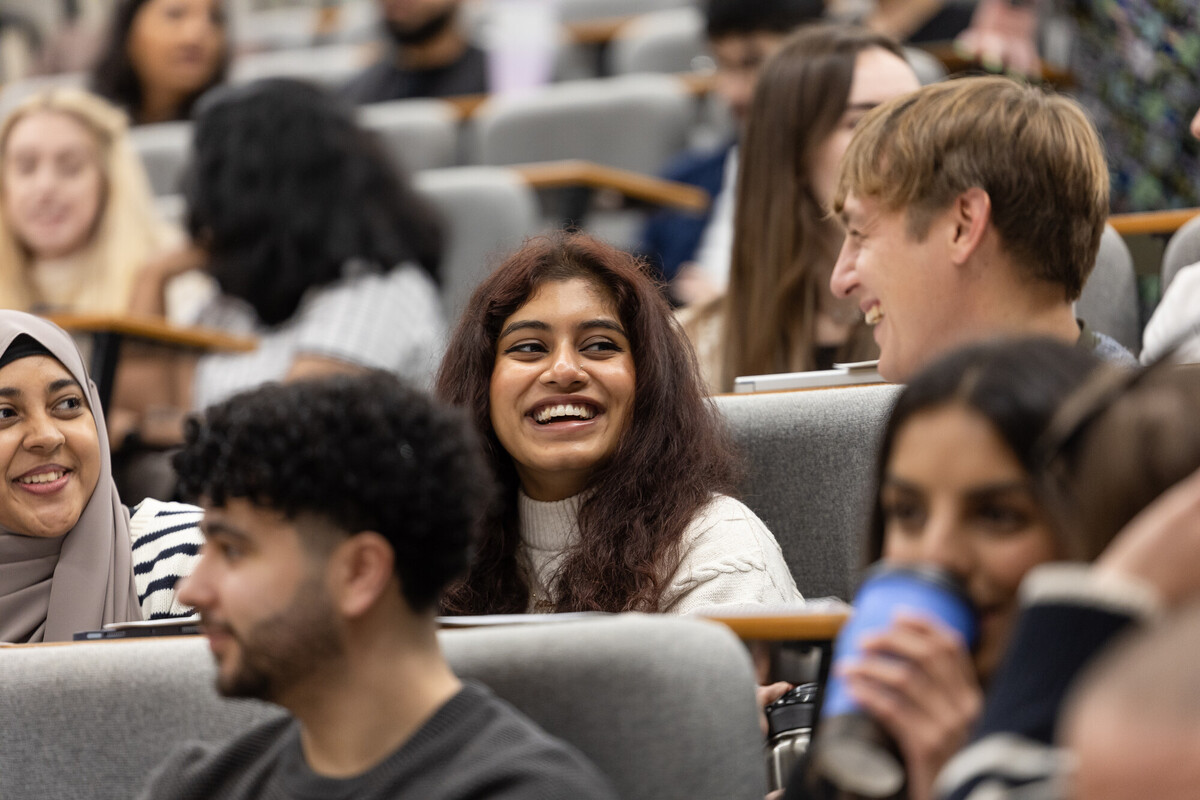 A smiling young woman with long dark hair sits in a tiered lecture hall surrounded by other Chester university students who are engaged and conversing.