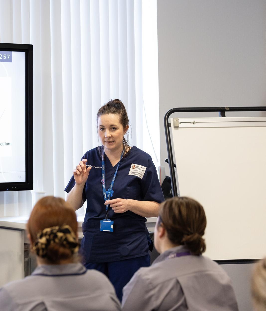 A healthcare training session, for midwifery students. A female instructor in navy blue scrubs is explaining a procedure.