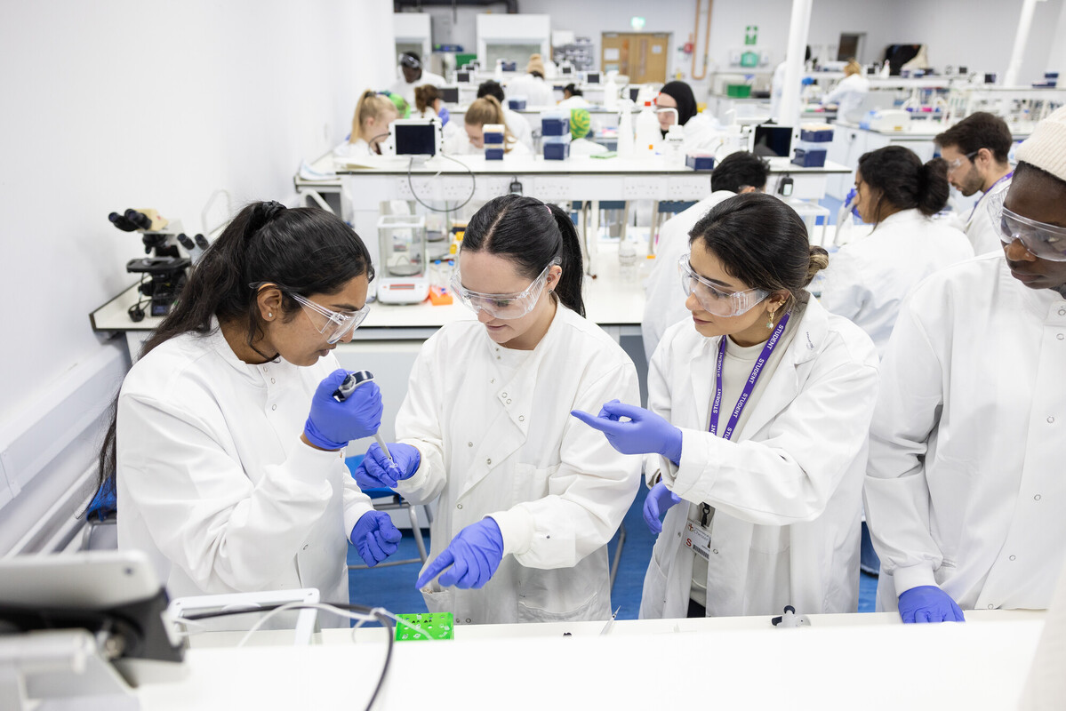 People wearing lab coats and safety goggles carry out a practical experiment at a laboratory bench in a busy teaching lab.