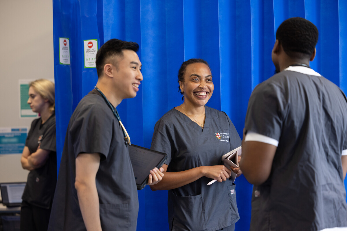 Chester medical students in grey uniforms chat and smile in front of blue curtains, reflecting a collaborative clinical learning environment.