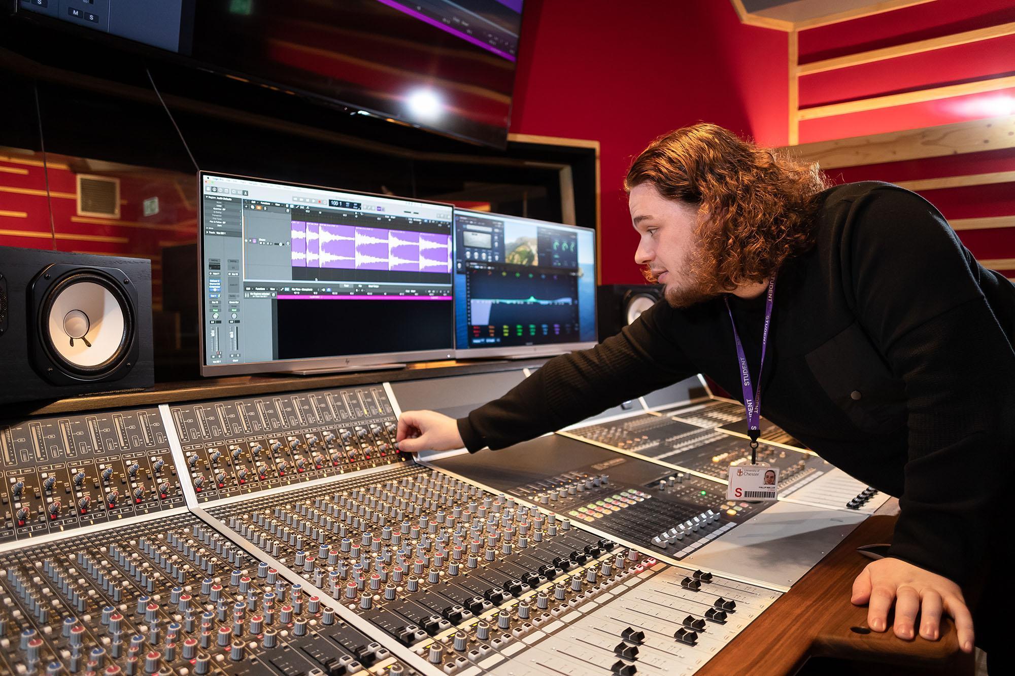 Music Production BA (Hons) student working on a professional mixing console in a recording studio, adjusting sound levels and editing audio on computer software.