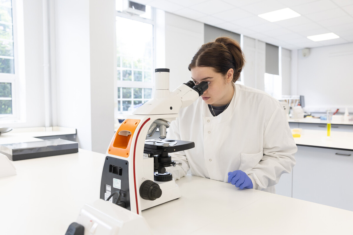 A student in a white lab coat and gloves examines a sample through a microscope in a bright, modern laboratory.