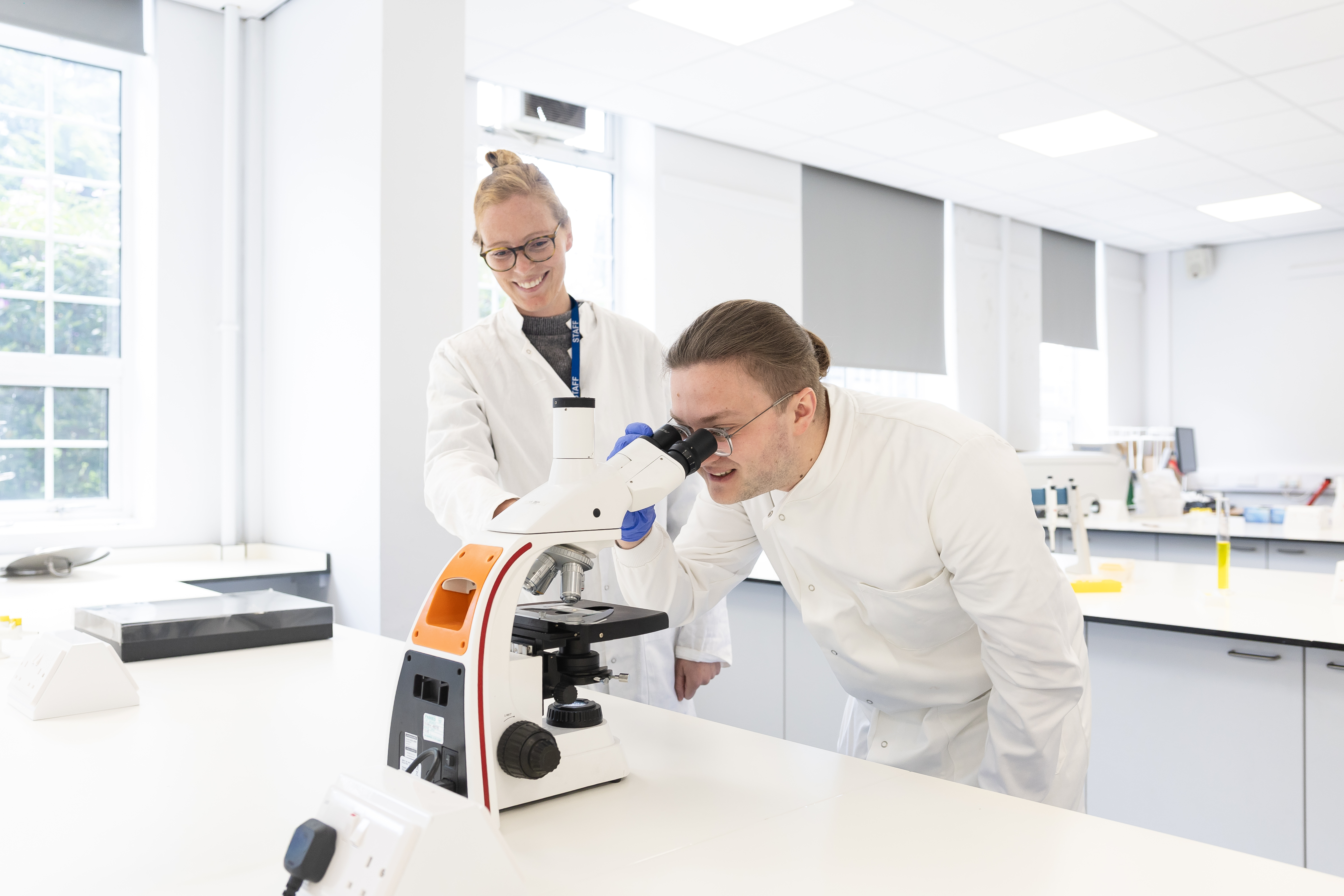 Two scientists in lab coats work in a bright lab, one using a microscope while the other observes.