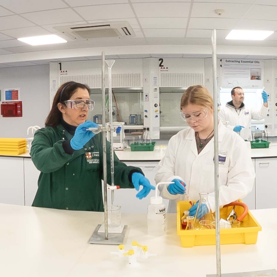 A laboratory scene where two students wearing safety goggles and gloves carry out a chemistry experiment with glassware and measuring equipment at a workstation