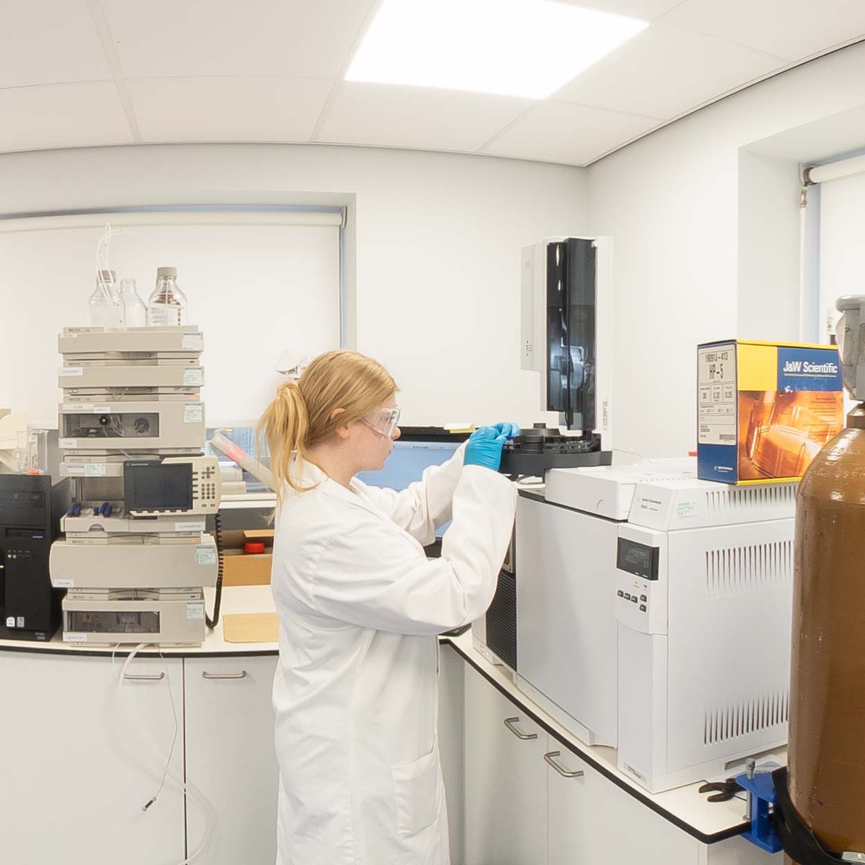 A student in a lab coat, gloves, and safety goggles operates analytical laboratory equipment on a workbench surrounded by scientific instruments.