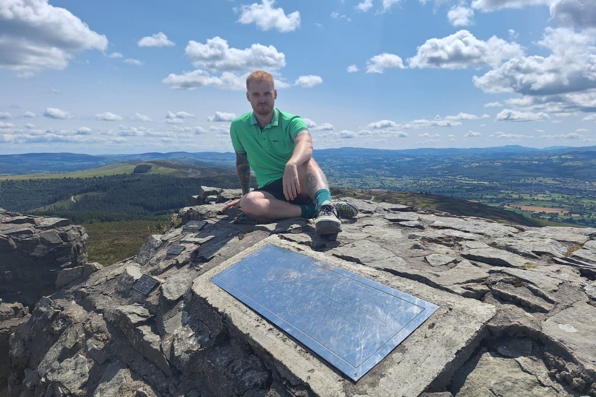 A man sitting on a rock at the top of a mountain, behind a plaque.