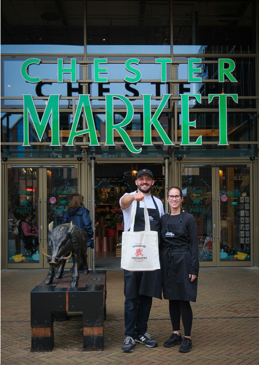 Two people, one holding a bag, smiling at the camera while standing outside Chester Market. The market signage is visible in green text. A bronzed wild boar is to the left of them as the market's symbol.