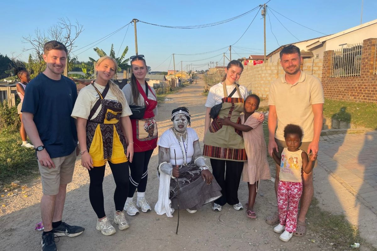 A group of five students looking at the camera, smiling, with two children and a person crouched down holding a stick. The group is on a path with a residential area in the background.