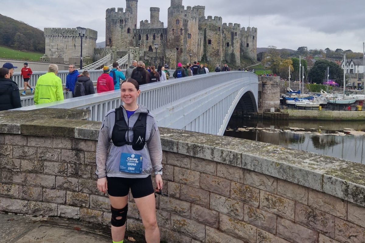 A woman in running gear and race number looking at the camera. In the background, runners going over a bridge over a river, watched by spectators. At the background is Conwy Castle.