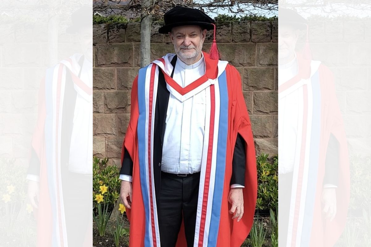 A man standing, looking at the camera, dressed in University ceremonial gown and hat. Behind him is a brown stone wall, flowers and a thin tree.