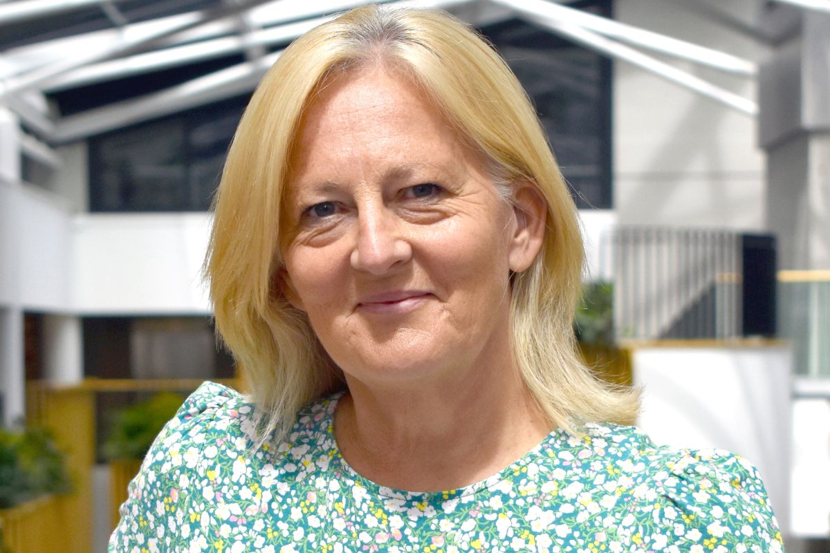 A head-and-shoulders photo of a woman in a green, yellow and white flowery dress looking at the camera. Out of focus in the background is a brightly lit modern room.