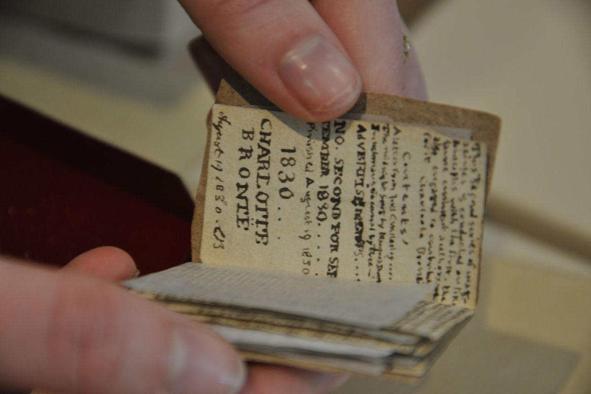 Thumb and fingertips leafing through a tiny manuscript. Among the very tiny text is 'Contents' and '1830 Charlotte Bronte'.