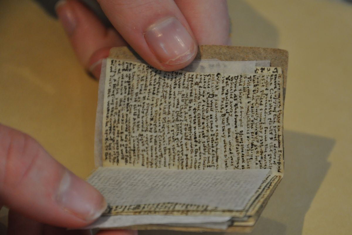 A tiny Charlotte Bronte manuscript with hundreds of words per page is held between two thumbs. The text is very hard to read.