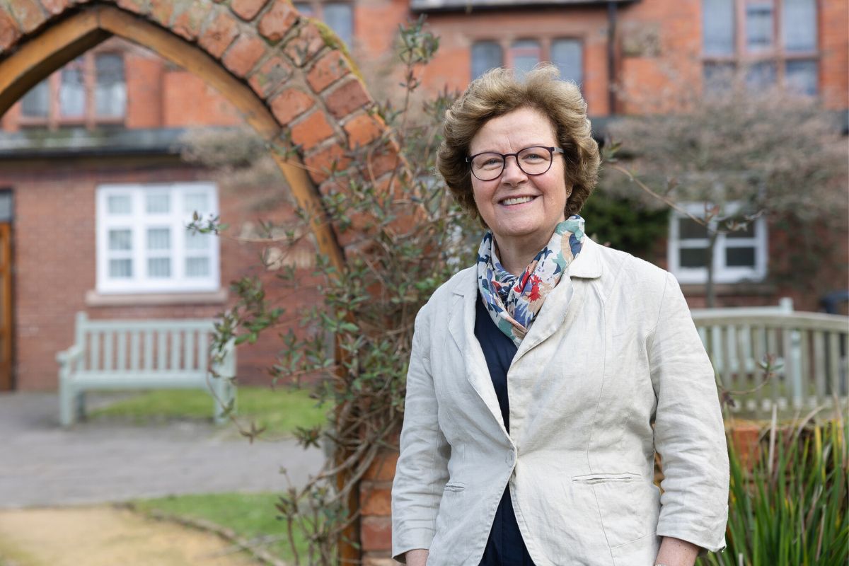 Vice-Chancellor Prof Eunice Simmons smiling while looking at the camera. In the background is a brickwork building, a walkthrough brick archway, benches, plants, grass and paths.