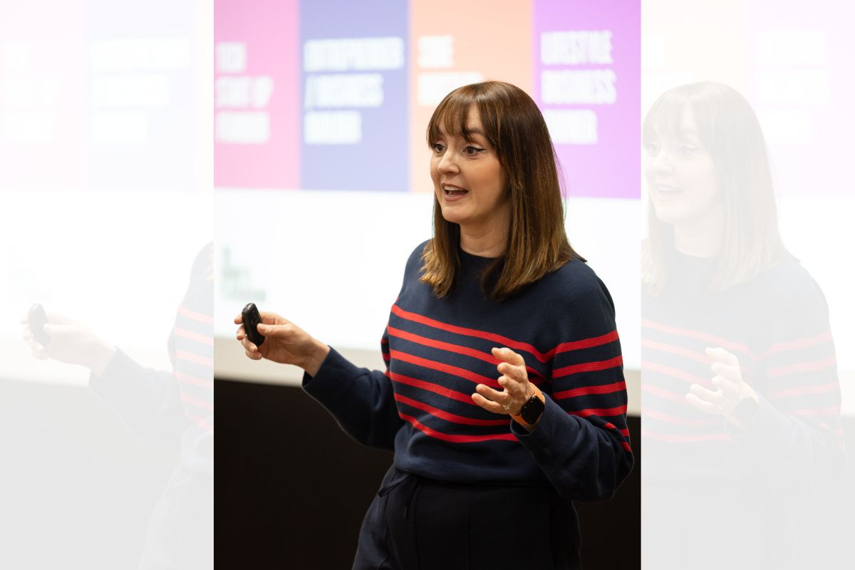 A woman giving a talk to an audience off-camera. In the background is a screen with some words which are out of focus.