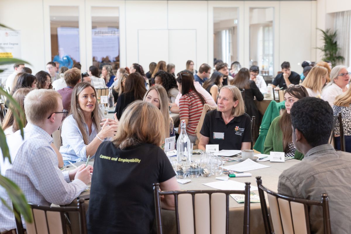 A busy group of people sat at round tables in discussions in a brightly lit room. One of the people as a T-shirt with the words 