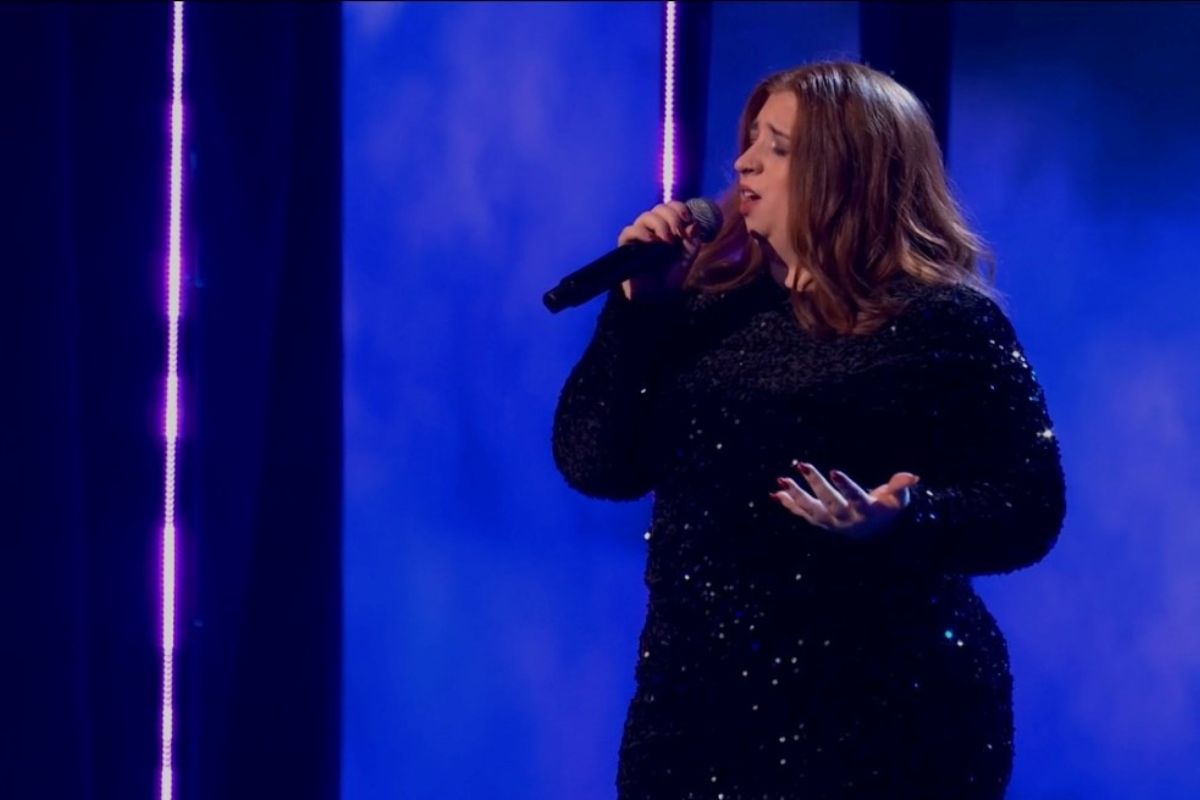 A woman in a glamorous dress with a microphone, in front of a dark blue stage backdrop.