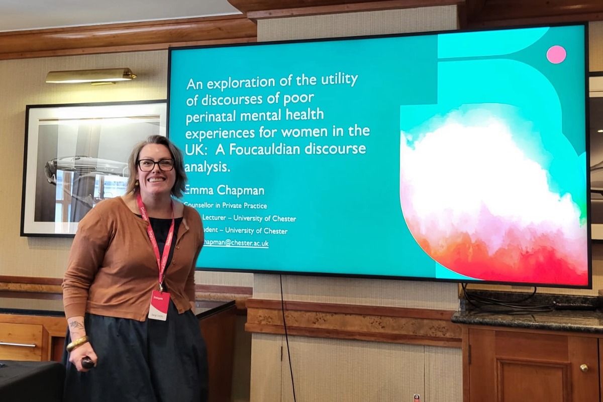 Emma Chapman standing and smiling at the camera. In the background is the title of her presentation on a big screen.