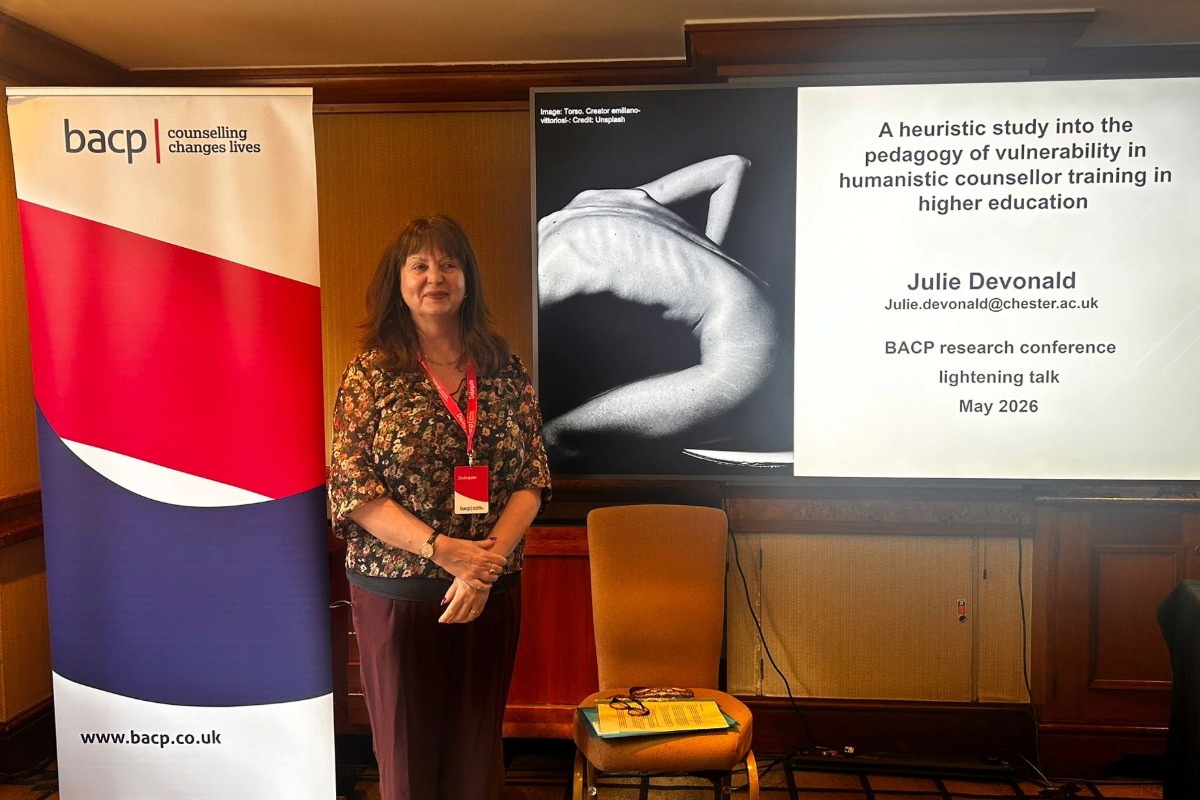 Julie Devonald standing and smiling at the camera. In the background is the title of her presentation on a big screen on the right, and the BACP logo on a stand on the left.