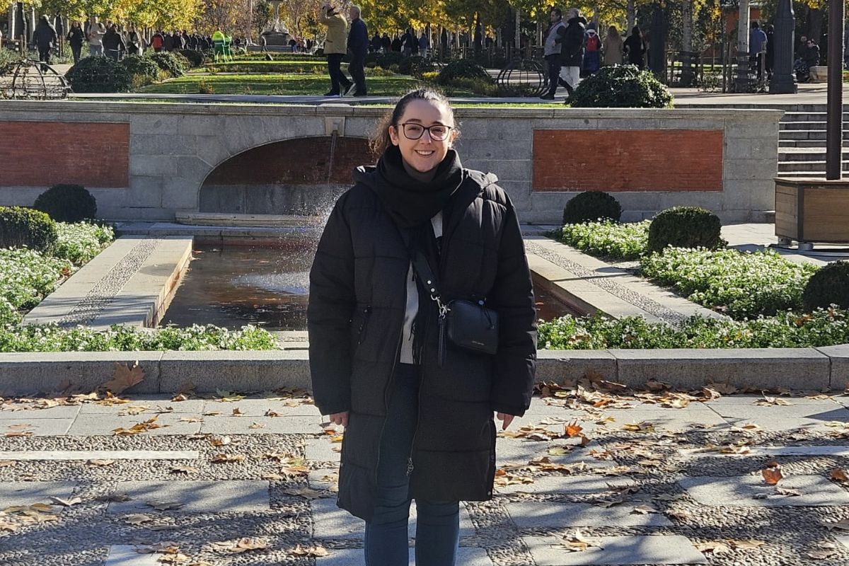 A woman in a coat standing and smiling at the camera in a picturesque courtyard garden area on a sunny day.