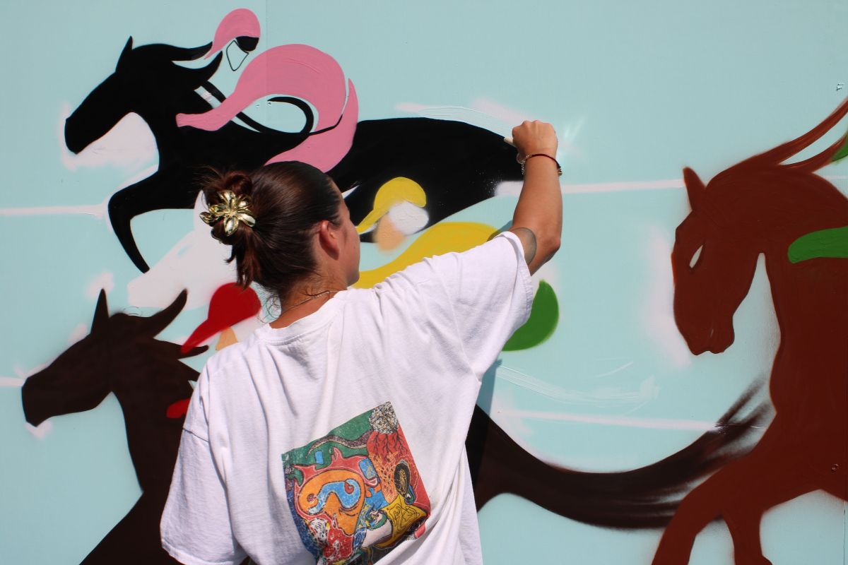 A woman with her back to camera, painting a mural on a wall. The mural is of jockeys in bright clothing on racehorses.